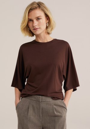 Blonde Frau mit kurzen Haaren, die goldene Creolen-Ohrringe trägt, ein dunkelbraunes lockeres Kurzarmoberteil und graue karierte Hosen mit Händen in den Taschen.