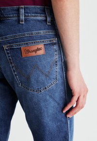 Tumši zilas džinsus ar ādas plāksteri, uz kura ir redzams "Wrangler" logo. Aizmugures kabatām ir unikāla dūriena zīmējums.