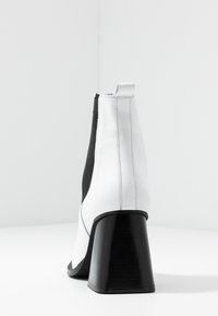Botín de tobillo blanco de piel sintética con un alto tacón negro de bloque, paneles laterales elásticos y un diseño elegante; costuras visibles en las uniones.