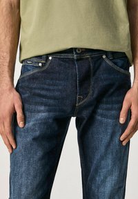 Tmavě modré džíny z denimu s přímým střihem, světlým šitím, pěti kapsami a kovovými knoflíky. Kombinovány s světle olivově zelenou košilí.