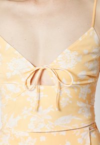 Vestido amarelo floral com decote em V, alças finas e detalhe de laço na frente, feito de um tecido liso e leve.