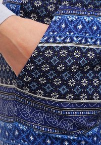 Blåmönstrat tyg med intrikata mönster, med geometriska former, blommotiv och varierande texturer. Ficka synlig på sidan.