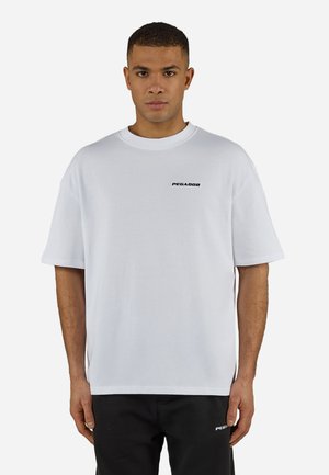 Άνδρας που φοράει λευκό πολύ μεγάλο μπλουζάκι με το λογότυπο "PEGADOR" στο στήθος και μαύρο παντελόνι με το ίδιο λογότυπο στο αριστερό μηρό, στέκεται μπροστά σε λευκό φόντο.