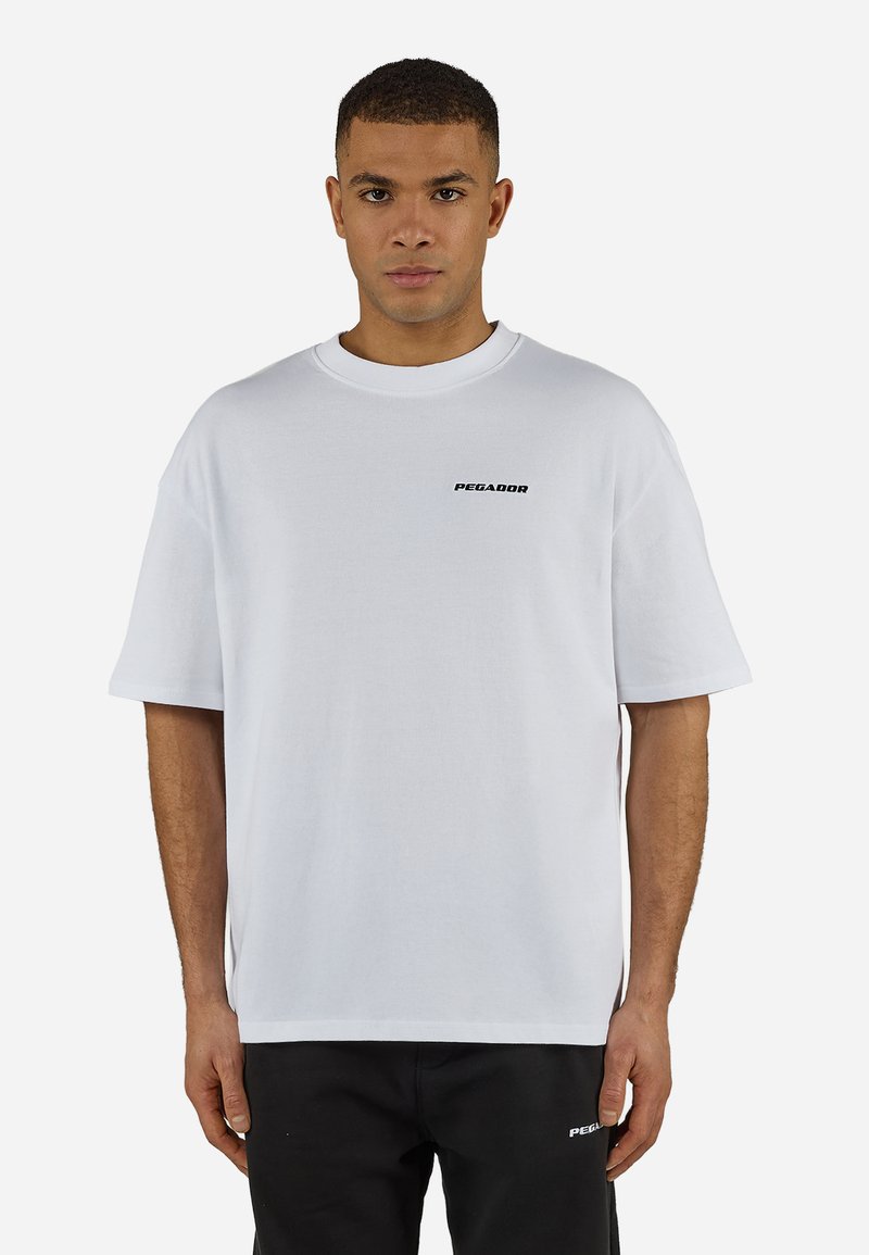 Vīrietis valkā baltu pārlielu t-kreklu ar uzrakstu "PEGADOR" uz krūtīm un melnas bikses ar atbilstošu logo kreisajā augšstilbā, stāvot pret baltu fonu.