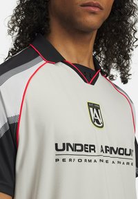 Šedé športové tričko s čiernymi akcentmi, červeným lemovaním a logom Under Armour. Má golierový dizajn a tkaninu odvádzajúcu vlhkosť.