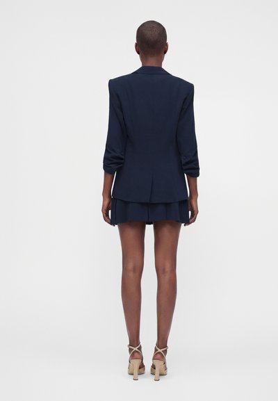Femme aux cheveux courts portant un blazer bleu marine et une jupe courte assortie, vue de dos, avec des sandales à talons beiges.
