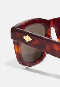 Skildpaddefarvede solbriller med en glat overflade, udstyret med brune tonede linser og et guld logo-akkent på siden. Firkantet rammedesign.