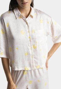 Chemisier à manches courtes, boutonné, en tissu léger de couleur rose pâle avec des motifs floraux jaunes. Présente une poche sur la partie gauche de la poitrine.