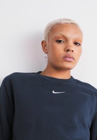 Marineblå hettegenser med rund hals og en liten hvit Nike-logo på brystet; myk stoff, avslappet passform, og lange ermer.