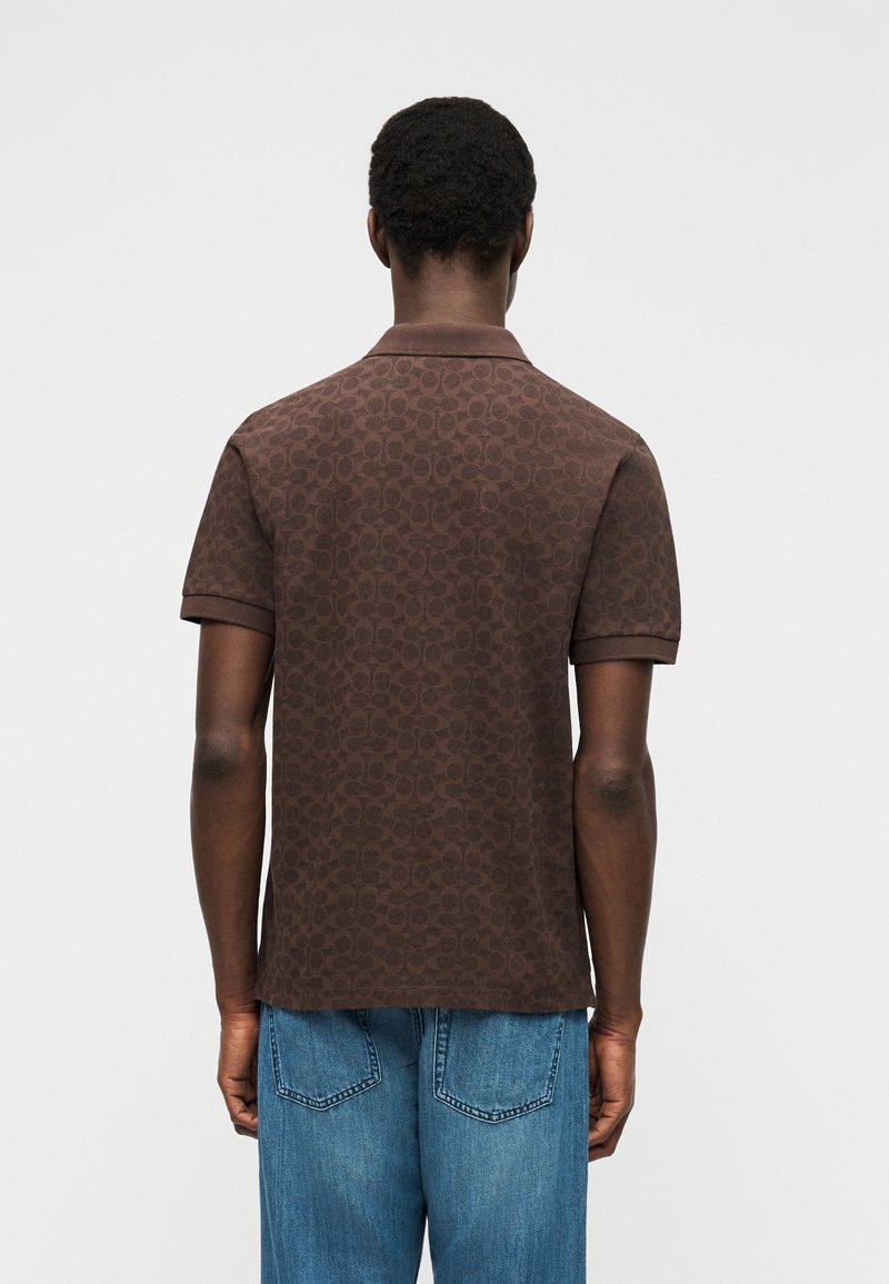 Man facing away wearing a brown patterned polo shirt and blue jeans against a plain gray background.