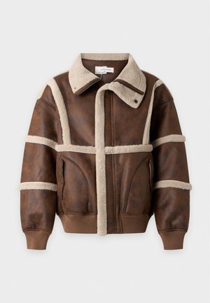 Un homme présente une veste marron doublée de peau lainée, une chemise blanche, un pantalon olive et des bottes noires sur un fond de studio uni.