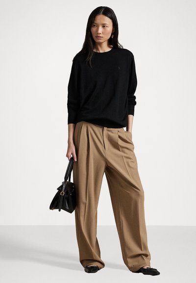 Black knitted sweater with long sleeves, relaxed fit. Tan wide-leg trousers with pleats. Model holds a small black bag, wearing black flats.