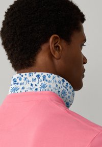 Homme aux cheveux courts et bouclés portant une chemise rose avec un col blanc affichant un imprimé tropical bleu de palmiers et de huttes, vu de dos.