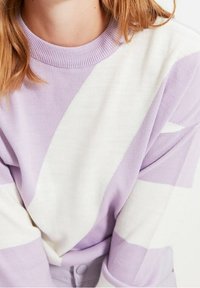 Detailní záběr na osobu, která má na sobě pletený svetr s levandulovými a bílými pruhy, s žebrovaným límcem a viditelnými světle hnědými vlasy.