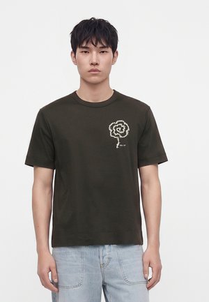 Jeune homme portant un t-shirt vert foncé avec un motif de fleur spirale blanche sur la poitrine et un jean bleu clair, debout devant un fond blanc.