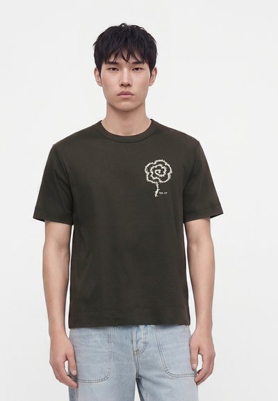 Νέος άνδρας που φοράει ένα σκουροπράσινο t-shirt με λευκό σχέδιο σπειροειδούς λουλουδιού στο στήθος και ανοιχτό μπλε τζιν, στέκεται μπροστά σε λευκό φόντο.