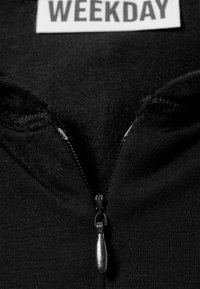 Sort lynlåstrøje med metallynlås og en tekstureret trækflig. Mærket viser "WEEKDAY." Stoffet fremstår glat og elastisk.
