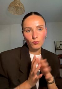 Junge Frau mit dunklen Haaren in einem braunen Blazer, die mit den Händen in einem gut beleuchteten Raum zeichnet, darüber ist eine geflochtene Leuchte sichtbar.
