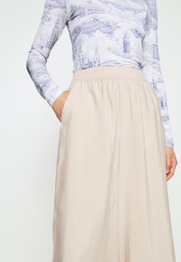 Femme portant un pantalon ample beige taille haute avec une ceinture élastique, et un haut ajusté à manches longues avec des motifs architecturaux bleus, main dans la poche.