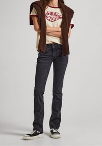 Jeans in denim nero con vestibilità slim, abbinati a una t-shirt crema con grafica rossa, e un maglione lavorato a maglia marrone drappeggiato sulle spalle.