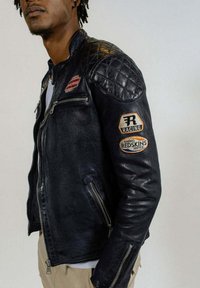 Homme portant une veste de course en cuir noir matelassé avec des poches zippées et des patchs sur la poitrine et le bras, debout devant un fond uni.