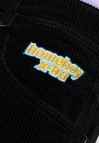 Černá kordurovina s vertikálním žebrováním, na níž je vyšitý text "homeboy x-tra" v oranžové a modré barvě. Text je stylizovaný a tučný.