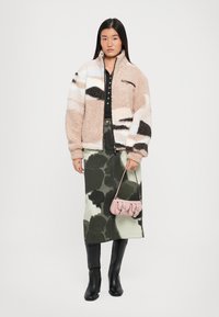 Jachetă din fleece bej cu modele negre și albe, purtată cu o fustă verde tie-dye și cizme negre până la genunchi. Accesoriile includ o poșetă roz.