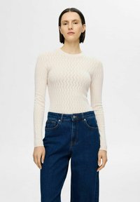 Ljus creamfärgad stickad topp med ett diamantramönster, långa ärmar, kombinerad med höga mörkblå jeans. Slät textur och figurnära design.