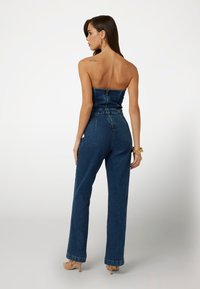 Džínový overal s nepriehľadným, priliehavým korzetom, so zipsom na zadnej strane, širokými nohavicami a tmavomodrou prateľnou farbou s minimálnymi kovovými detailmi.