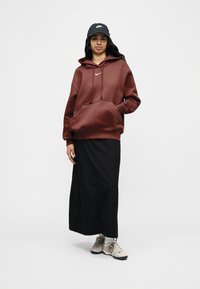 Γ υναίκα που φοράει σκούρα κόκκινη μπλούζα με κουκούλα Nike, μακριά μαύρη φούστα, γκρι καπέλο Nike, λευκές κάλτσες και ανοιχτόχρωμα αθλητικά παπούτσια, στέκεται με το ένα χέρι στην τσέπη.