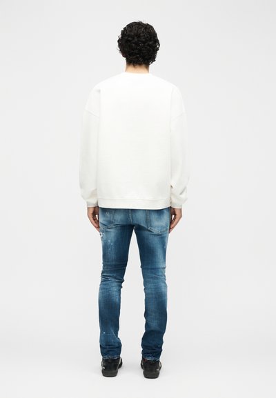 Άτομο με σγουρά μαλλιά, που στέκεται στραμμένο προς τα πίσω, φορώντας λευκό πουκάμισο με μακριά μανίκια, μπλε ξεθωριασμένο τζιν και μαύρα παπούτσια πάνω σε μονόχρωμο φόντο.