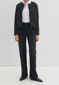 Veste courte noire texturée portée sur une chemise blanche, associée à un jean noir taille haute et des chaussures noires pointues. Design simple et structuré.