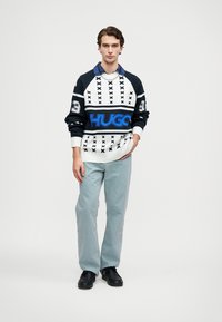 Pleteni pulover v črno-belih barvah z modrim logotipom "HUGO", 3D vzorec in rebrastimi manšetami. Usklajen s svetlo modrimi jeans hlačami in črnimi čevlji.