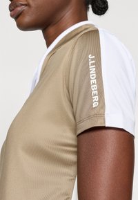 Kratke rokave športna majica v tan in belem blagu. Ima logo "J.LINDEBERG" v beli barvi na rami. Gladka tekstura.
