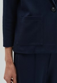 Marineblazer aus strukturiertem Stoff, mit einer einzigen Vordertasche, einem Knopfverschluss und einem geraden Saum über maßgeschneiderten Hosen.