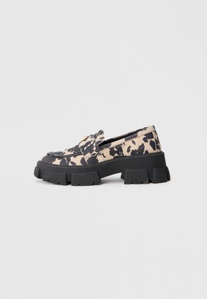 Loafer mit klobiger Sohle, beigem und schwarzem Animal-Print-Obermaterial und goldfarbenem Metalldetail auf dem Spann vor hellgrauem Hintergrund.
