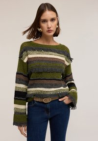 Pulover tricotat în dungi, de culoare verde, maro și crem. Oferă un material texturat, tiv cu ciucuri și mâneci late. Asortat cu blugi de culoare bleu închis și o curea.