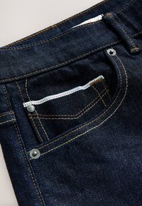 Donkerblauwe denimstof met een voorzak met koperen klinknagels, contrasterende oranje stiksels en een klein binnenzakje met witte rand.