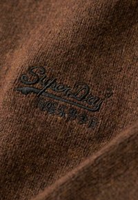Tessuto in maglia marrone con logo e testo ricamati in nero "SuperDry" sulla superficie, che mostra la texture e il dettaglio ravvicinato.