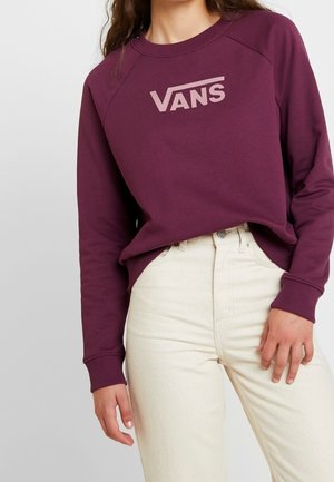 Personne portant un sweat à capuche Vans bordeaux à manches longues et un jean taille haute couleur blanc cassé, debout avec une main détendue sur le côté.