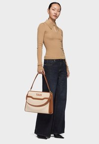 Camiseta beige de manga larga de punto con diseño de ojo de cerradura, combinada con vaqueros anchos de pierna oscura. Bolso estructurado marrón y crema con el logo "TOUS".