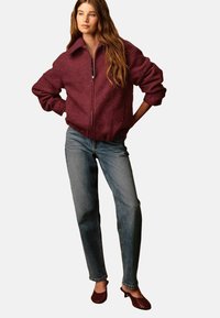 Femme portant une veste zippée bordeaux, un jean droit bleu et des escarpins à talons kitten bordeaux, se tenant avec les mains sur les hanches.