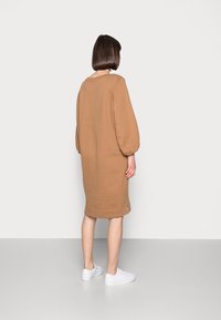 Hnědá, oversized mikinová šaty s balónovými rukávy, žebrovaným lemem a bočními rozparky; v kombinaci s bílými teniskami na světle šedé podlaze.