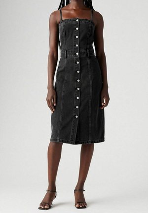 Frau trägt ein schwarzes, ärmelloses Denim-Midikleid mit Knopfleiste und dünnen Trägern sowie schwarze Sandalen mit offenem Zehenbereich und Absatz vor einem schlichten Hintergrund.