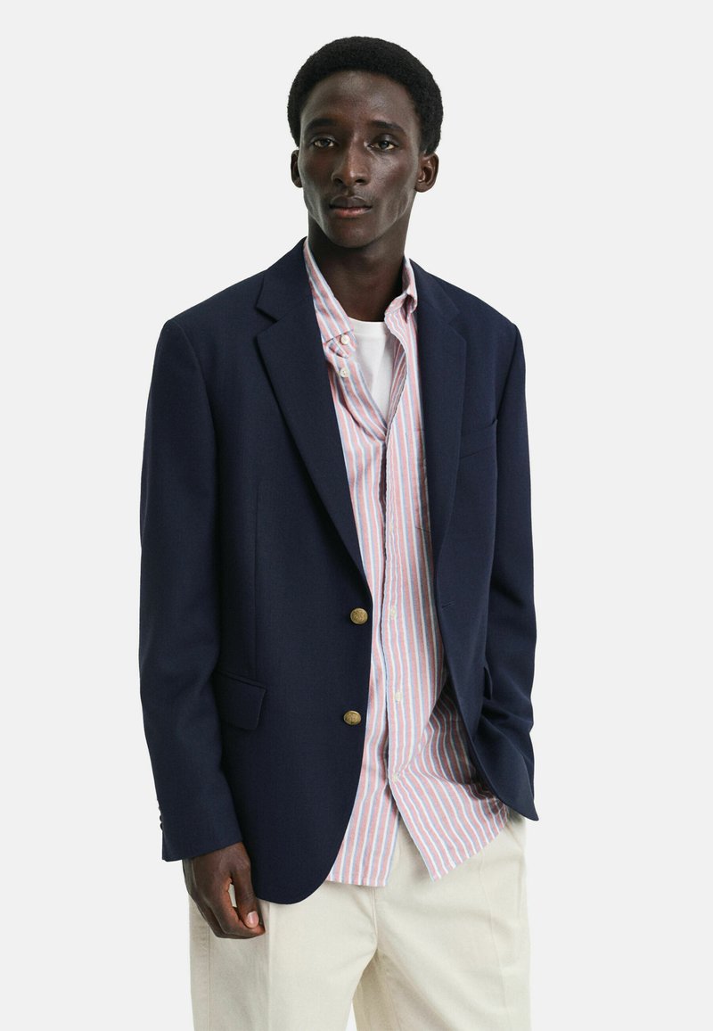 Blazer navy con bottoni dorati, sovrapposto a una camicia a righe rosa chiaro e bianca. Il modello indossa pantaloni color crema, dal design minimalista.