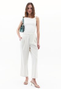 Witte getailleerde tanktop, crème hoge taille broek met zijzakken en sandalen met gouden banden met hak. Kleine groene handtas accentueert de look.