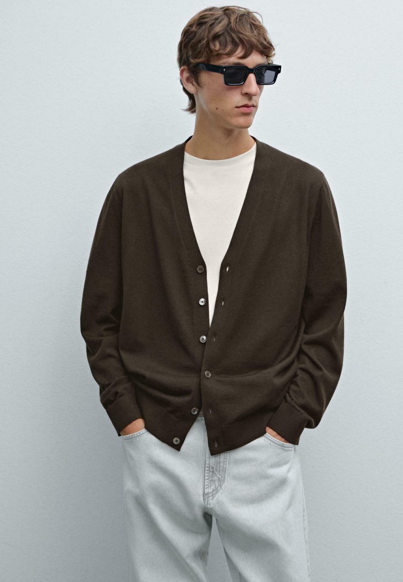 Brun kofta med V-ringning och fem knappar, kombinerad med en vit skjorta och ljusblå jeans. Modellen bär svarta solglasögon.
