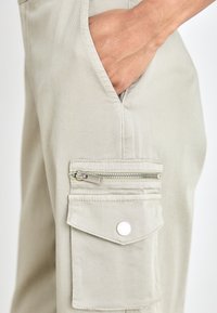 Beige cargo pants in durable fabric with a side pocket featuring a zipper and flap, and a standard front pocket for added utility.
