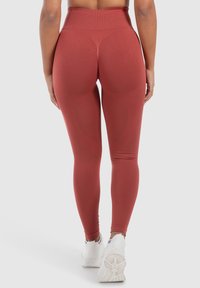 Leggings taille haute, ajustés, de couleur rouille avec une texture lisse, présentant des détails de contournage subtils et portés avec des chaussures de sport blanches.