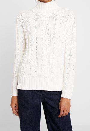 Pull à col roulé en maille torsadée blanche porté avec un jean en denim bleu foncé, montrant la partie supérieure du corps et les bras d'une personne.